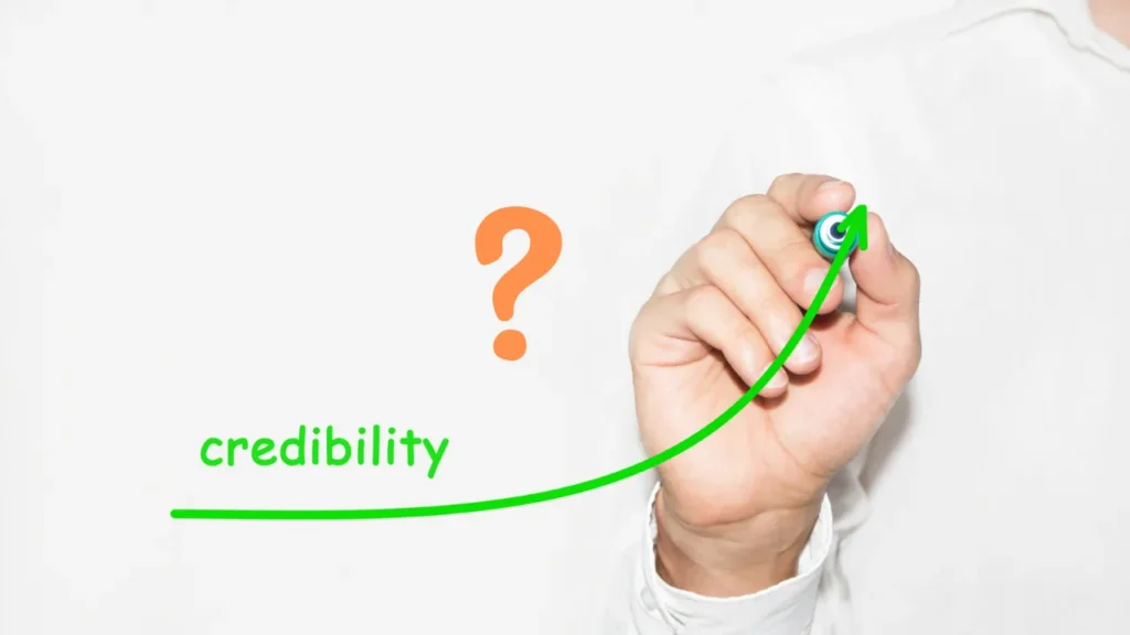 An individual's hand drawing an ascending green line graph with a marker, symbolizing growth in credibility. To the left, an orange question mark represents inquiry or doubt. The word 'credibility' is written in green at the bottom of the graph's trajectory, suggesting a direct correlation between the inquiry and the concept of credibility. The background is plain white, focusing attention on the central imagery. This visual metaphor emphasizes the importance of credibility in professional settings, relevant to the blog post titled 'Is Your Website Making You Look Unprofessional?'.